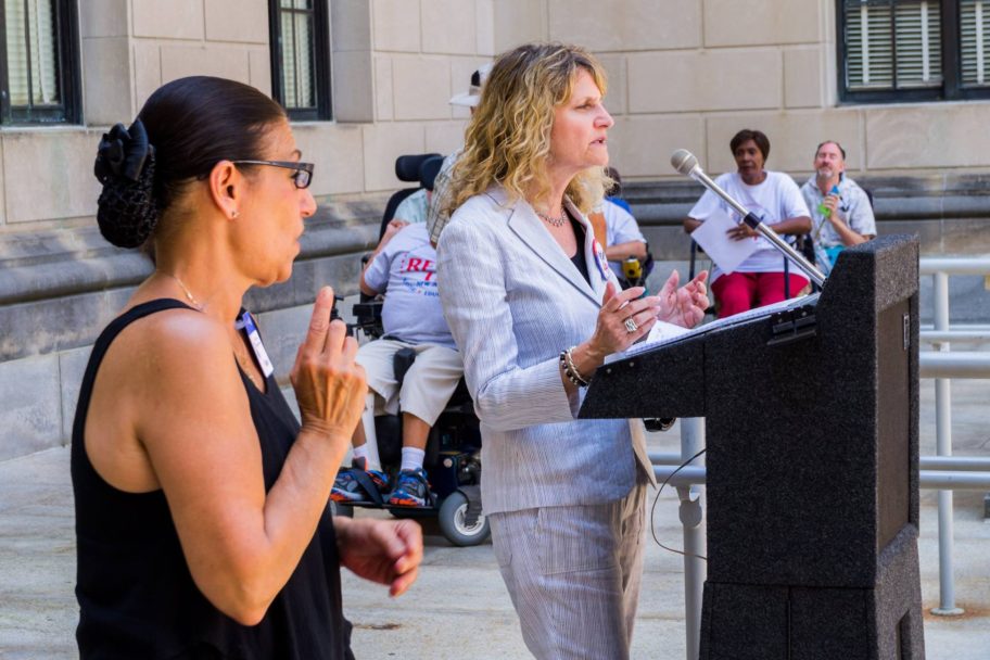 female in business suit standing at podium outside with a sign language translator beside her speaking to a group of people outside