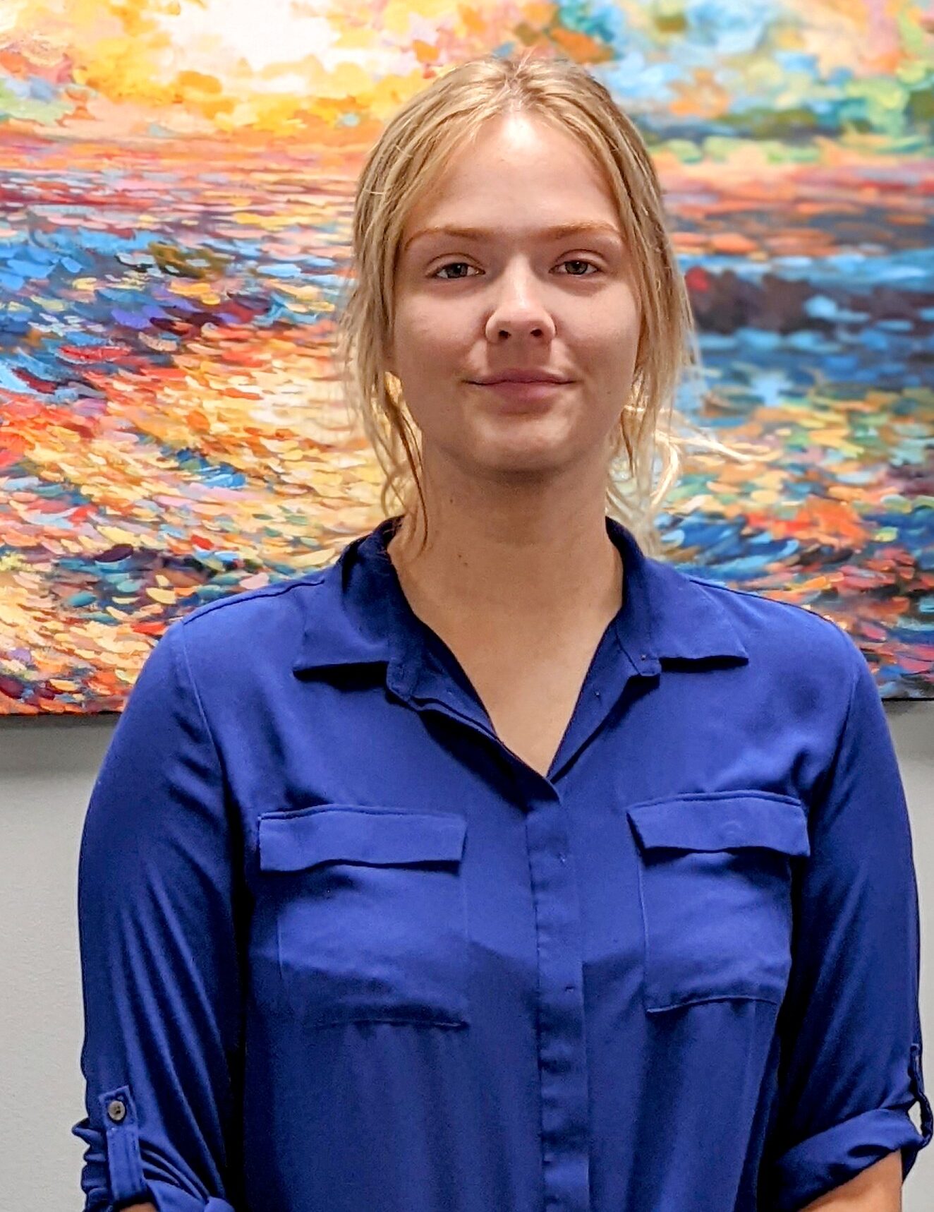 White female with blonde hair wearing a blue button up shirt and standing in front of a colorful abstract painting smiling at t he camera