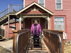 Independence, Inc. consumer wearing a bright pink jacket, black pants and top, uses a walker to navigate down her new entry ramp. Her apartment in a large, ornate, pink home with many stairs, is in the background.