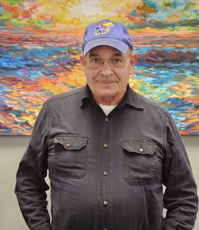 Older male wearing a blue Kansas University hat, glasses and a gray long sleeve shirt, standing in front of a colorful painting