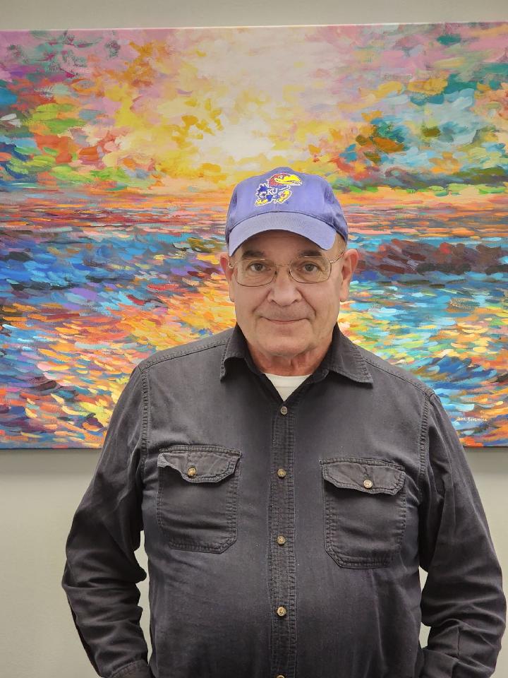 Older male wearing a blue Kansas University hat, glasses and a gray long sleeve shirt, standing in front of a colorful painting