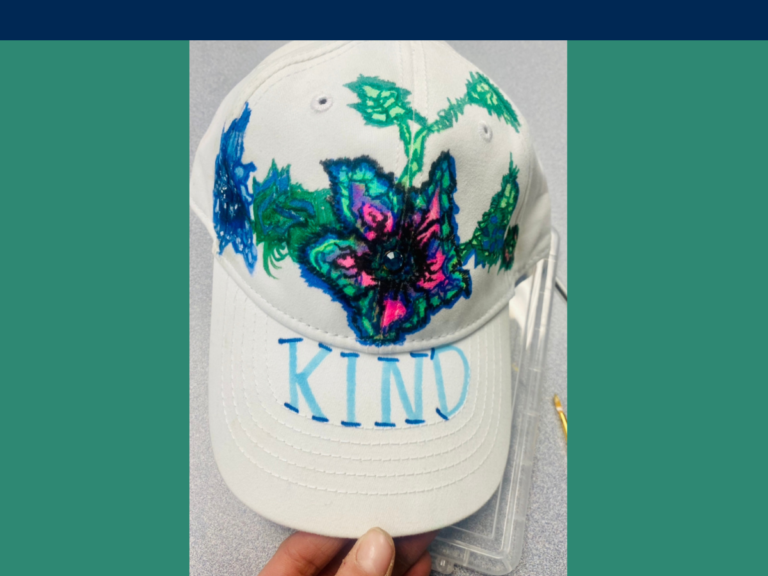 fingers holding the brim of a white hat with hand painted flowers and the  word KIND