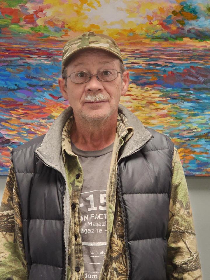 Older white male with a gray mustache wearing a camo style hat, camo shirt and dark gray vest, standing in front of a colorful painting