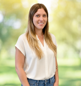 Posed staff photo of a young woman with long dark blonde hair wearing a white top and blue jeans, smiling at the camera