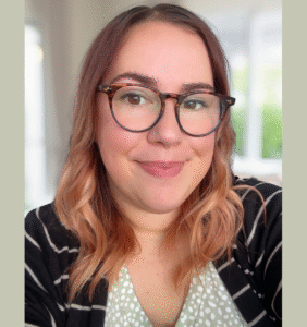 Female with reddish brown hair and dark framed eye glasses, smiles at the camera