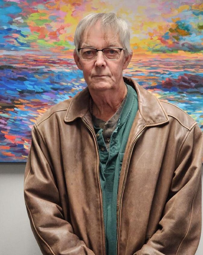 Older white male with short gray hair wearing a brown jacket and glasses and standing in front of a colorful painting