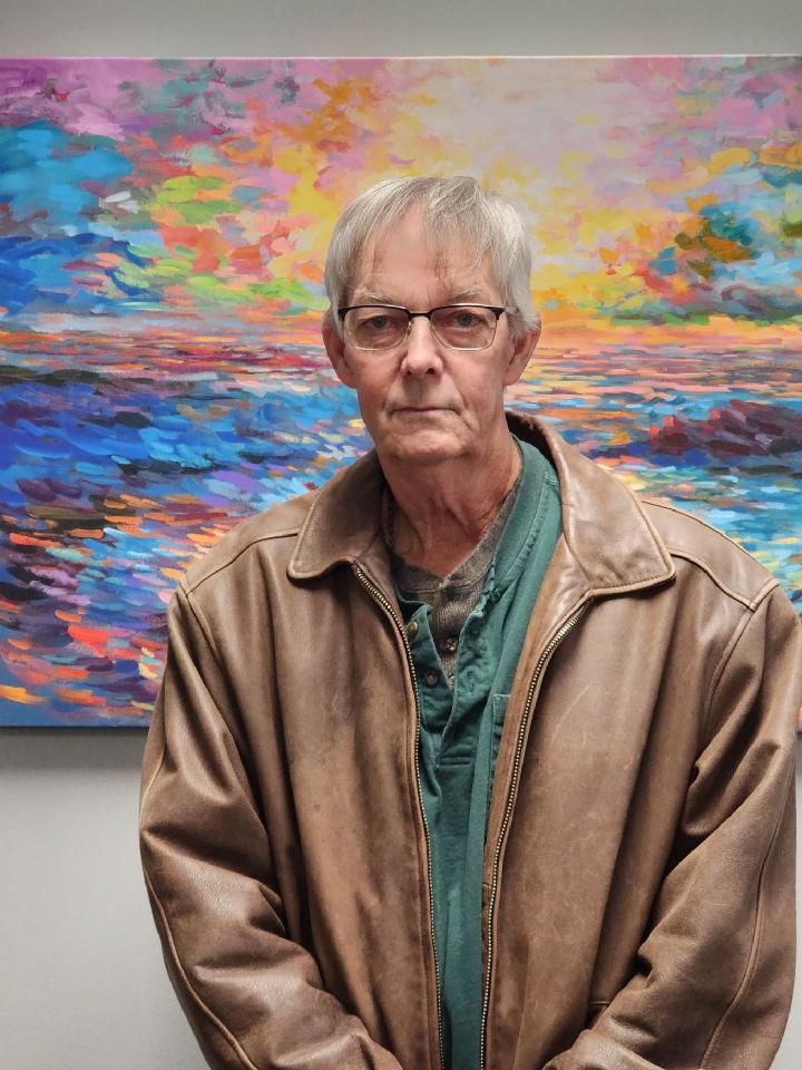 Older white male with short gray hair wearing a brown jacket and glasses and standing in front of a colorful painting