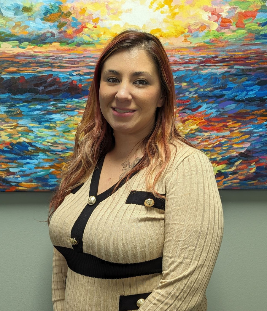 Female with long reddish brown hair standing in front of a color abstract painting, smiling at the camera