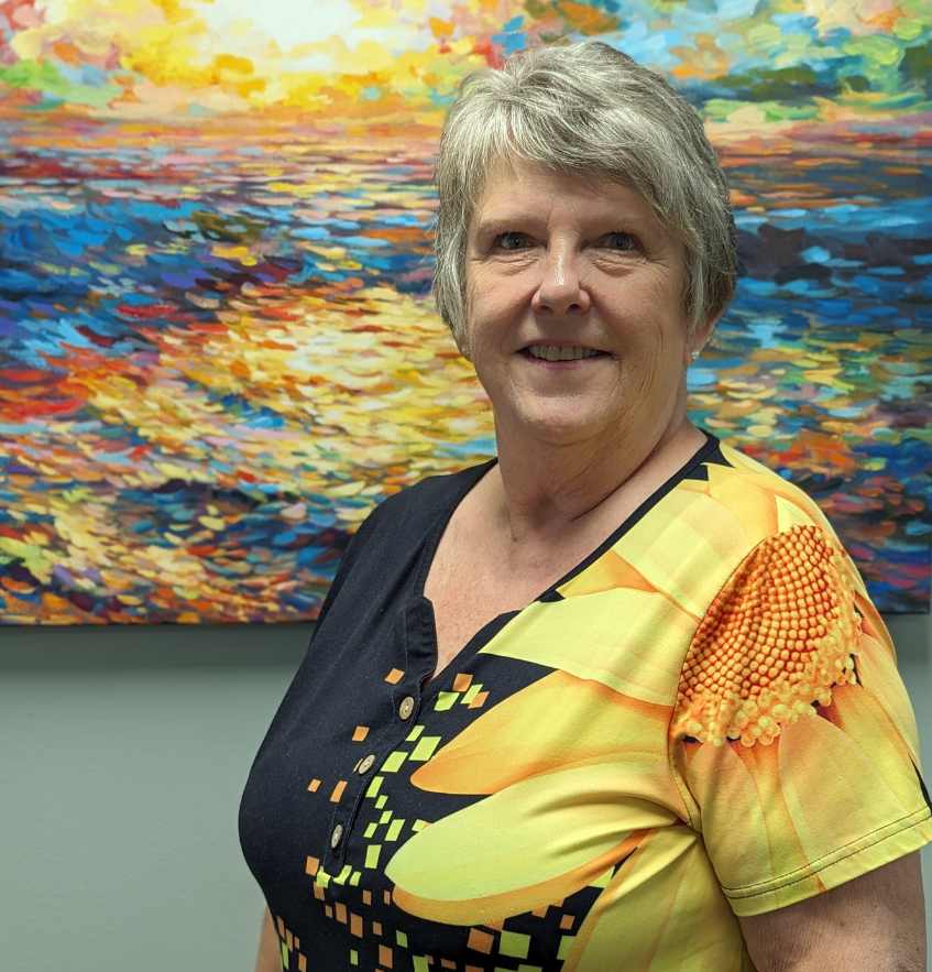 Female with short gray hair wearing a colorful blouse with a sunflower and bright yellow, green and orange dots with black background, looking at the camera and smiling