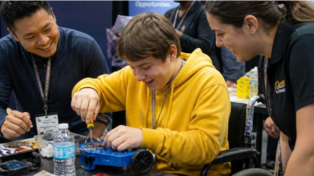 A teenage boy with medium brown hair wearing a bright yellow hoodie, sits at a table using a scredriver to work on a mechanical box with two adults supervising
