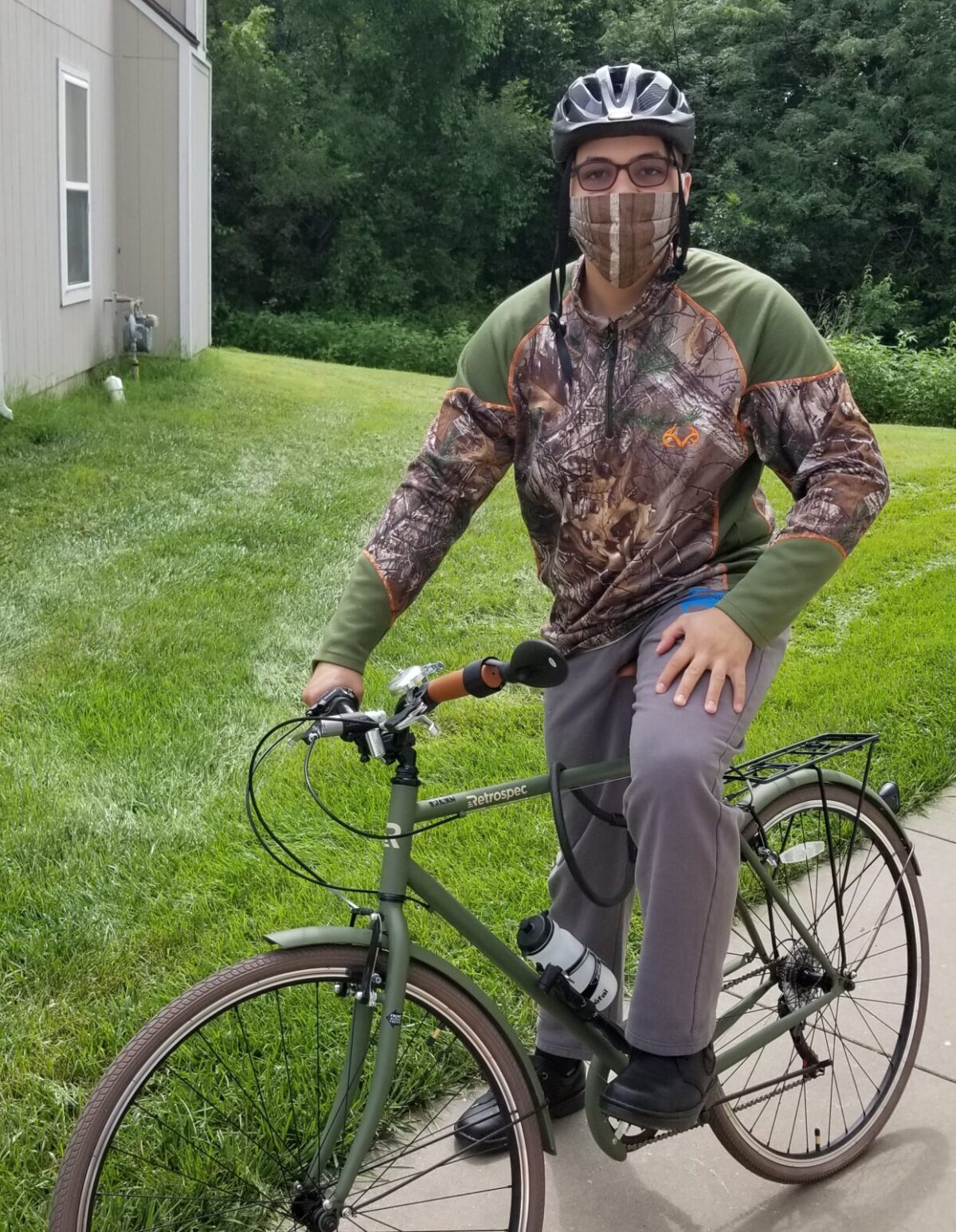 Independence, Inc. consumer poses on a sidewalk in front of his apartment, sitting on his bike wearing camouflage pattern jacket and gray sweats, wearing a bike helmet .