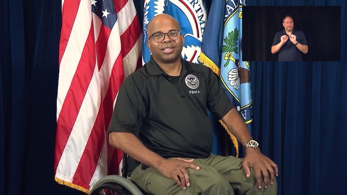 Sherman Gillums, Jr with FEMA Office of Disability Integration and Coordination sitting on a stage in his wheelchair in front of an American flag, looking at the camera