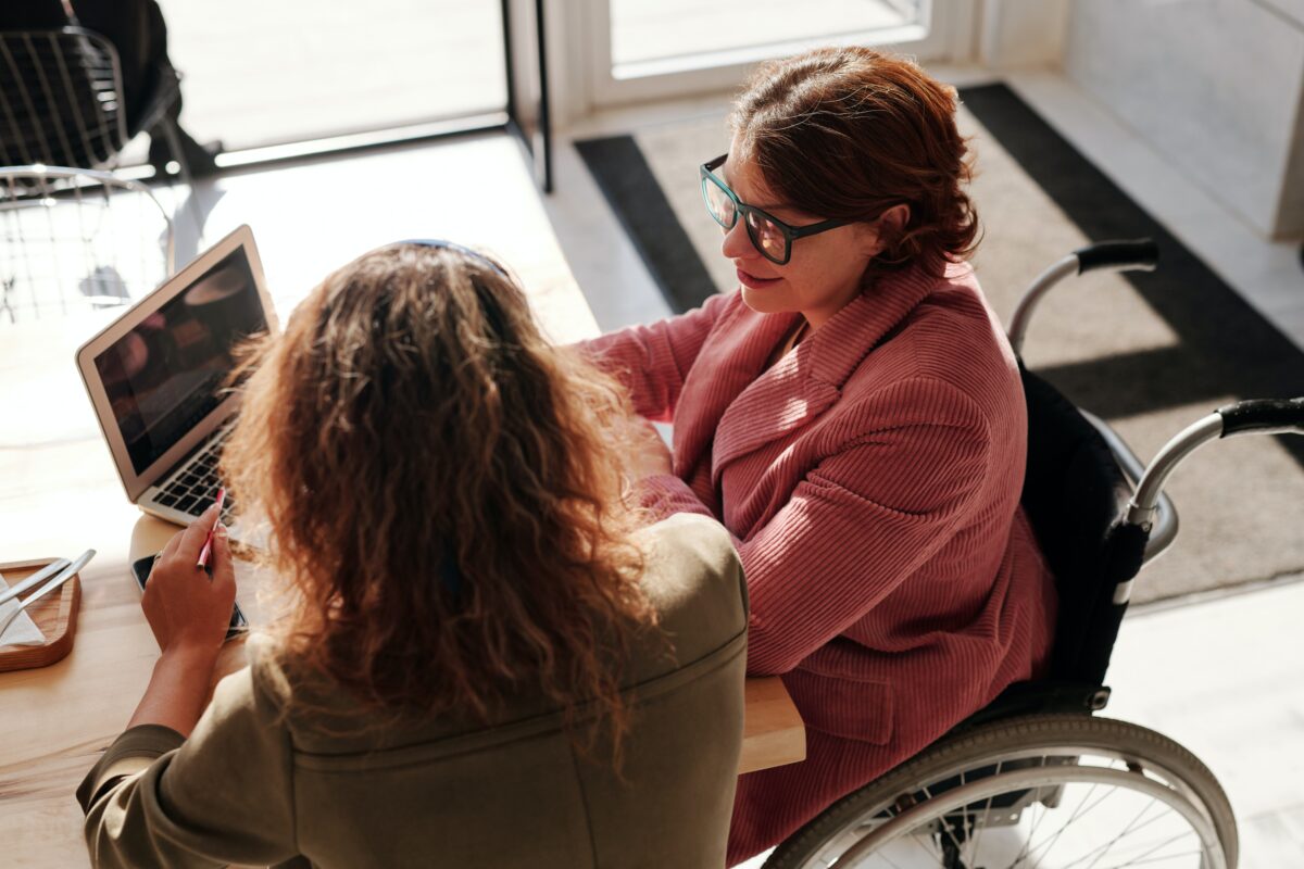 female with brown hair and glasses sitting in a wheelchair at a desk with another female with longer reddish brown hair, back turned to the camera, assisting with a project