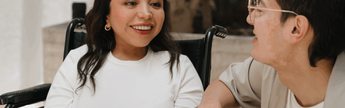 Two young adults, a female with medium skin tone and long dark hair sitting in a wheel chair and a male with medium skin tone with short dark hair and glasses, sit in an outdoor plaza style area near a large building.