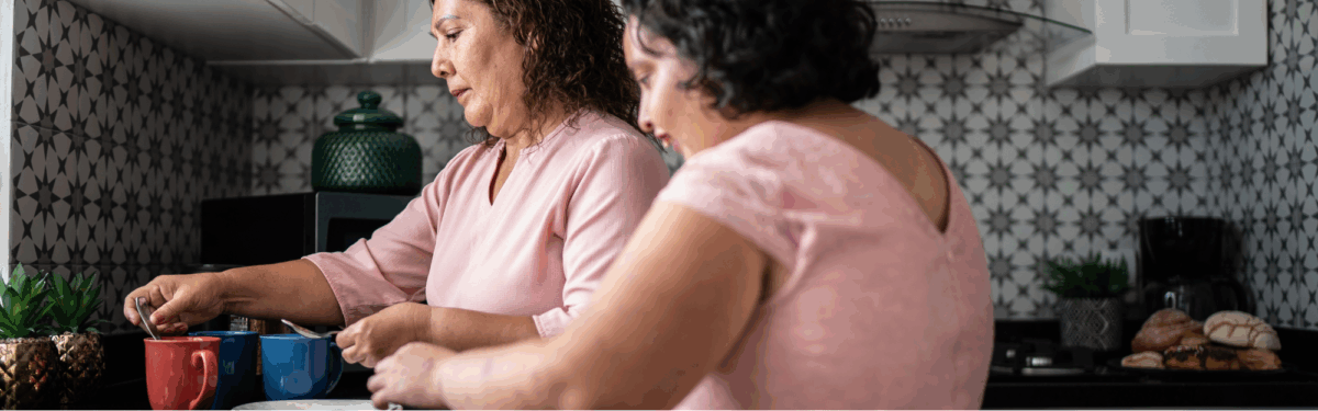 Two women are in a kitchen, leaning against a counter with a sink in front of them. Sunlight coming in through the window.