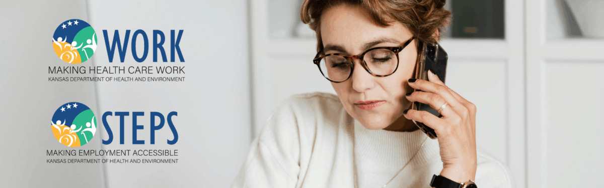 A female with short brown hair wearing brown glasses and a white shirt, is sitting at a desk holding a phone to her ear.