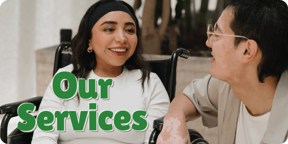 Two young adults, a female with medium skin tone and long dark hair sitting in a wheel chair and a male with medium skin tone with short dark hair and glasses, sit in an outdoor plaza style area near a large building.